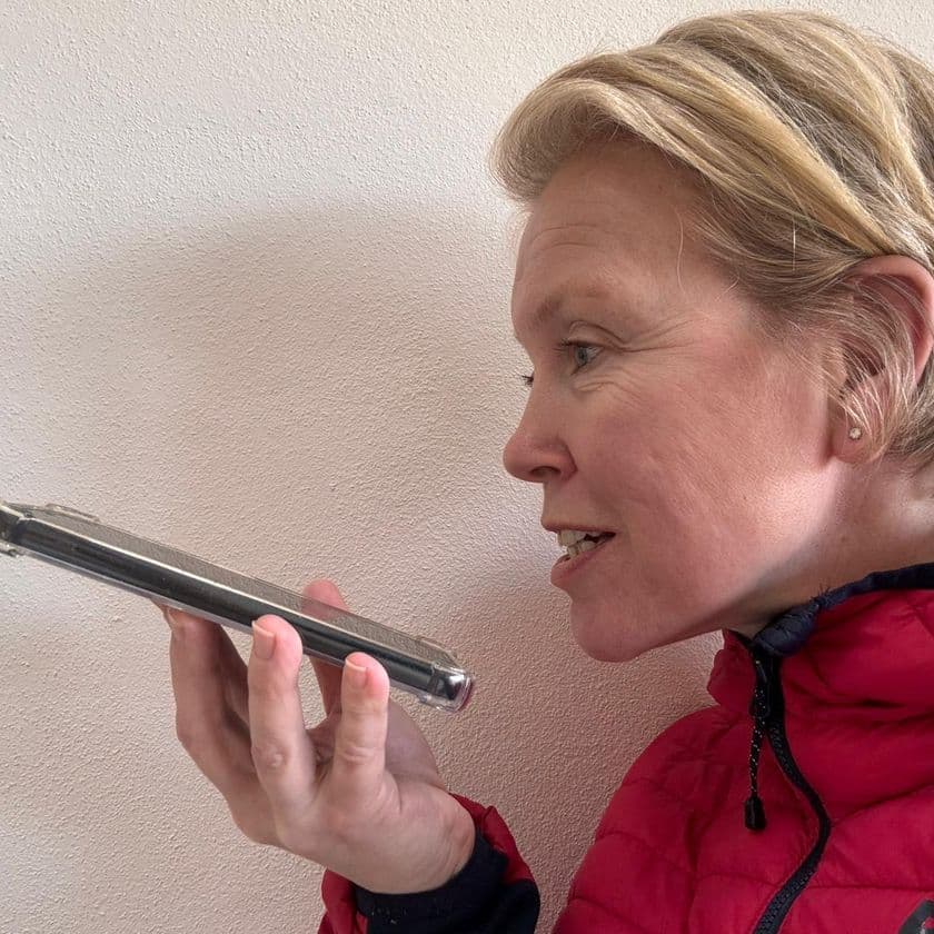 Person in a red jacket holds a smartphone horizontally, speaking into it closely, against a plain wall background.