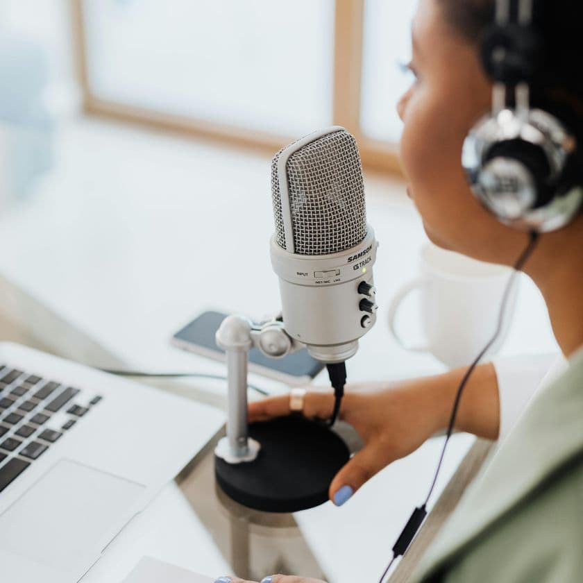 Person wearing headphones speaking into a microphone, sitting at a desk with a laptop and a coffee cup in the background.