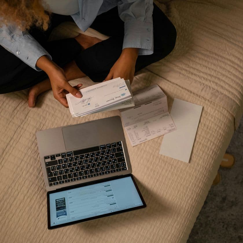 Person sitting on bed with a laptop and holding papers, possibly checks, while managing finances or completing tasks.