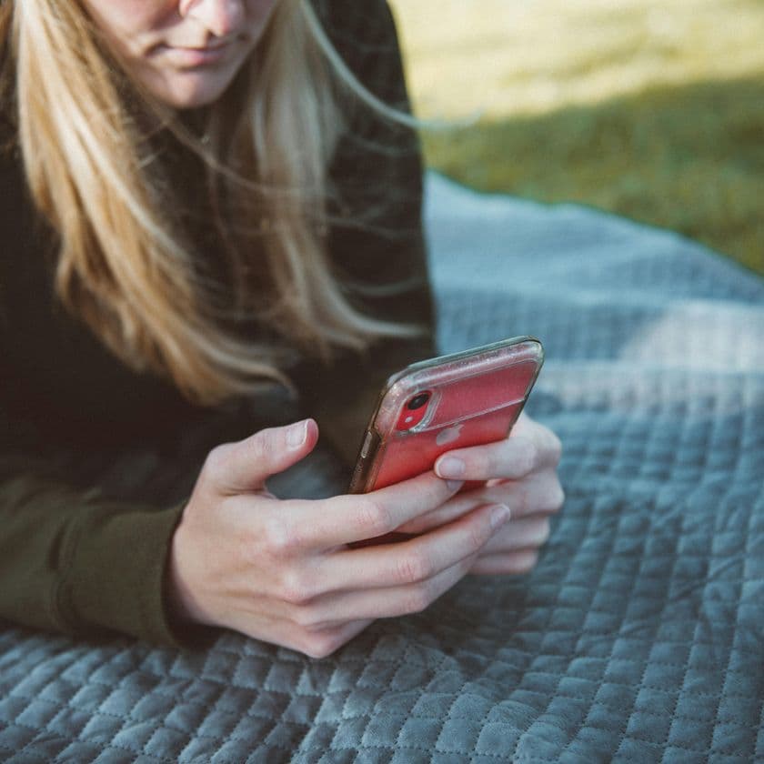 Person lying on a gray blanket outdoors, holding a red phone, with long hair partially covering their face.